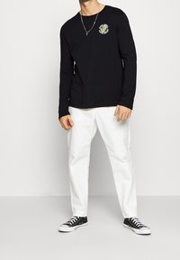 Zwart T-shirt met lange mouwen en een kleurrijk tijgerembleem op de borst, gecombineerd met witte broeken en zwarte sneakers. Platte textuur en casual pasvorm.