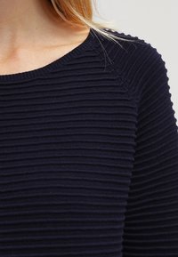 Maglione a coste navy con scollo rotondo ampio e superficie testurizzata. Il tessuto è morbido e presenta sottili linee orizzontali.
