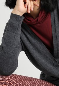 Grå cardigan med V-ringning, knäppning och ribbade ärmslut, lagd över en vinröd polokrage tröja, tillsammans med mönstrade leggings.