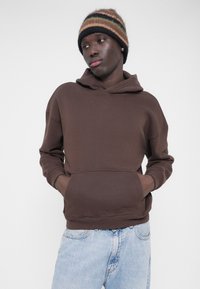 Καφέ φούτερ με κουκούλα και τσέπη κανγκουρό, με άνετη γραμμή, συνδυασμένο με τζιν σε ανοιχτό μπλε χρώμα. Το μοντέλο φοράει ριγέ πλεκτό σκουφάκι.