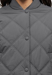 Γκρι εξωτερικό ένδυμα με επένδυση, που διαθέτει ψηλό γιακά, μεγάλες κουμπιά και ραφές διαμαντένιου σχήματος, κατασκευασμένο από λεία, ελαφριά υλικά.