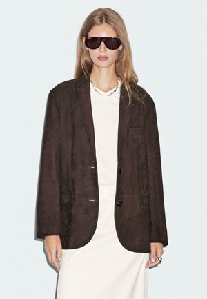 Brown suede blazer with a relaxed fit, featuring two front pockets, black buttons, and worn over a light-colored top. Model wearing sunglasses.
