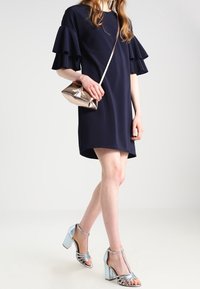 Marineblaues Kleid mit Rüschensleeves, metallische silberne Clutch und hellblaue High-Heelsandalen mit Riemchendetails und einem Blockabsatz.