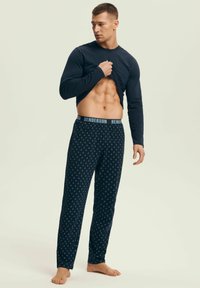 Mężczyzna w ciemnej, długiej koszuli podnoszący ją, aby pokazać wyrzeźbione mięśnie brzucha, w wzorzystych granatowych spodniach piżamowych z gumką "HENDERSON", bez butów.