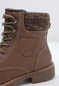 Botte montante en cuir marron avec bord en tricot, œillets métalliques, semelle texturée et détails cousus sur un fond uni.