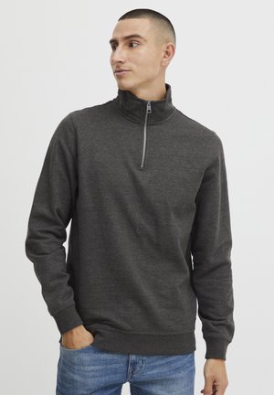 Sudadera de cuello con cremallera en gris oscuro, de manga larga, textura suave, con puños y dobladillo acanalados, combinada con jeans azules. Diseño simple y minimalista.