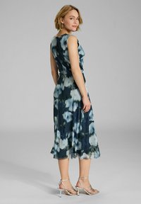 Ärmelloses Blumenkleid in Blau- und Grüntönen, fließender Stoff, knielang, mit tailliertem Schnitt und ausgestellt Rock. Silberne Riemchensandalen.