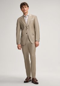 Beige Anzug mit maßgeschneidertem Design, ausgestattet mit zwei Knöpfen, Reverskragen und seitlichen Taschen, kombiniert mit einem gemusterten Hemd und braunen Schuhen.