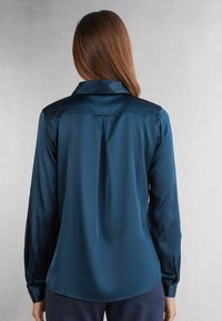 Saténová blůza ve tmavě tyrkysové barvě s dlouhými rukávy, límečkem a designem s sklady na zadní straně, který zdůrazňuje hladkou texturu a elegantní tvar.