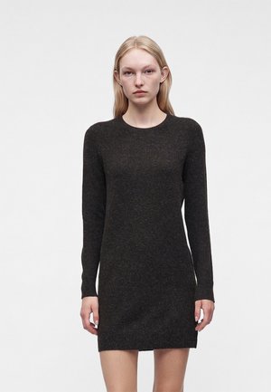 Femme blonde aux cheveux raides portant une robe moulante en maille gris foncé à manches longues, debout devant un fond blanc uni.