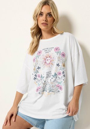 Kobieta nosząca białą oversize koszulkę z słońcem, kwiatami, motylami i napisem „Znajdź mnie tam, gdzie świeci słońce.”
