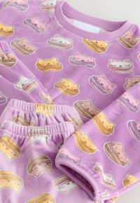 Lilla pyjama komplekt, mis on valmistatud pehmest kangast, millel on korduv muster kasside nägudest ja kihilistest kookidest. Pükste peal on elastne vöökoht.