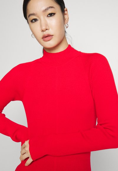 Pull à col roulé rouge côtelé avec des manches longues, présentant un design ajusté et un style raglan sans coutures. Texture douce sans motifs visibles.