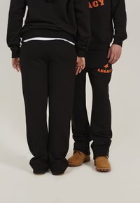 Haut à capuche noir et pantalon de survêtement, avec un texte "LEGACY" orange et un design d'étoile. Tissu doux, coupe décontractée et style casual.