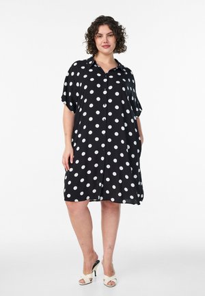 Kvinde med krøllet hår iført en løs sort kjole med hvide prikker og hvide åbne tå-sko, stående med hænderne i lommerne.