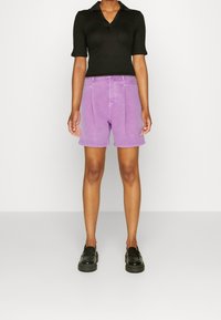 Top corto negro de manga corta con canalé y cuello, combinado con pantalones cortos de mezclilla morados de cintura alta. Unos zapatos de plataforma negros completan el conjunto.