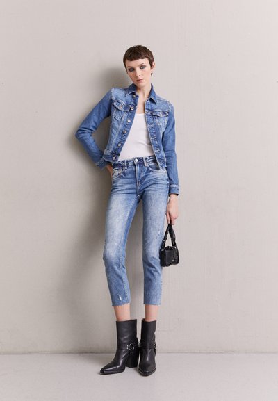 Veste en denim bleu clair avec poches avant, portée sur un débardeur blanc. Jean bleu clair déchiré et bottines noires à boucles complètent le look.