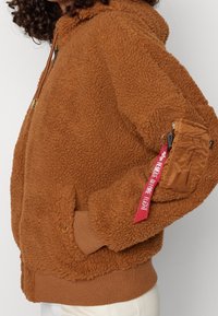 Hnědá fleecová mikina s texturovaným povrchem, plným zipem a žebrovanými manžetami. Obsahuje boční kapsu s červeným popruhem.