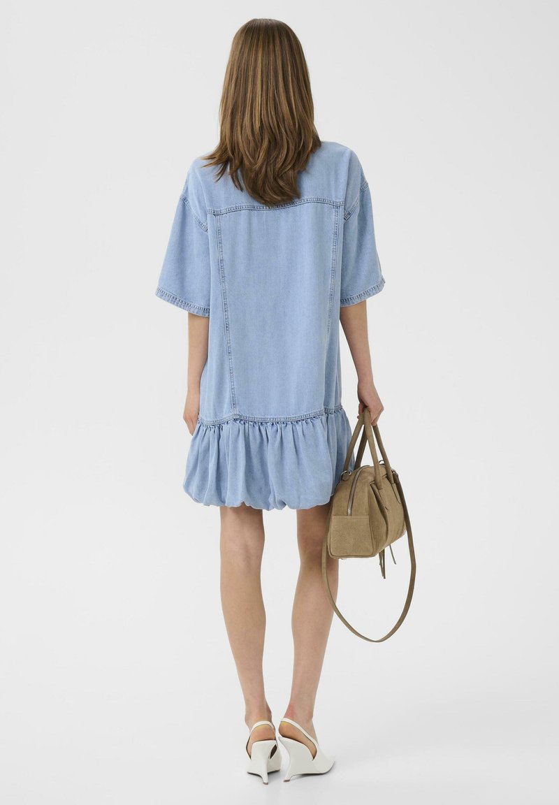 Vrouw met schouderlang bruin haar, gekleed in een loszittende lichtblauwe spijkerjurk en witte hakken, met een beige suède handtas naast zich.