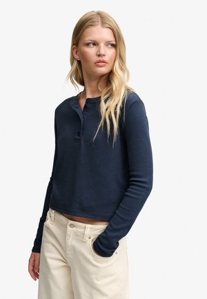 Lyshåret kvinde iført en navy langærmet henley-top og cremefarvede højtaljede bukser med den ene hånd i lommen, kiggende sidelæns.