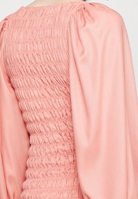 Vaaleanpunainen teksturoitu paita, jossa on rypytetty yksityiskohta vartalon ympärillä ja suurta huoliteltua hihaa, mikä korostaa pehmeää, joustavaa kangaspainatusta.