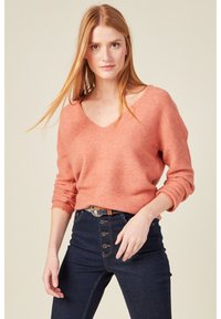 Korallfarbener V-Ausschnitt-Pullover mit langen Ärmeln, strukturiertem Strick, kombiniert mit dunklen, hoch taillierten Jeans mit Knopfdetails und einem Gürtel.
