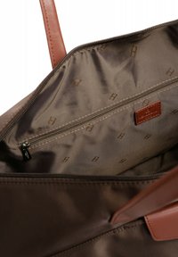 Interno di una tote bag in tessuto marrone con texture liscia, dotata di tasca con zip e un patch con logo. Manici in pelle marrone chiaro. Motivo logo sottile.