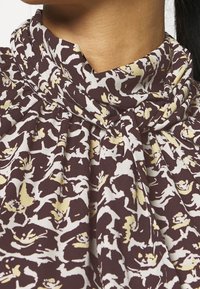 Braunblümchenbluse mit einem drapierten Ausschnitt. Der Stoff hat Creme- und Gelbtöne, die eine texturierte Optik erzeugen.
