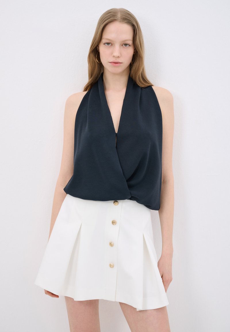 Top senza maniche di colore blu navy con un design drappeggiato, abbinato a una gonna plissettata bianca con bottoni dorati lungo il davanti.