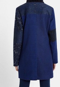 Marinblå kappa med ett texturerat rutmönster, kontrasterande paljettärmar och en strukturerad design. Kragen är mjuk och mörk.