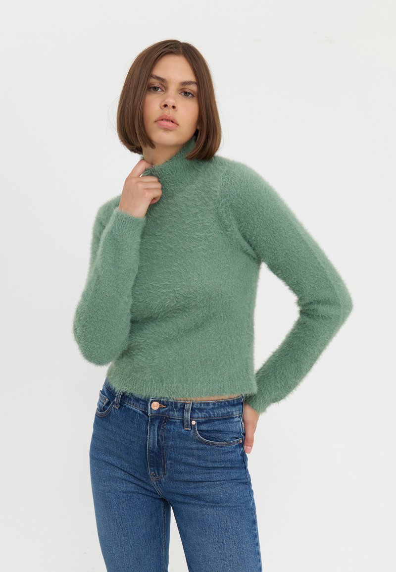 Maglione verde fluffy a collo alto con maniche lunghe, design aderente e polsini a coste, abbinato a jeans blu a vita alta.