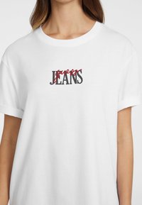 T-shirt bianco a maniche corte con testo nero e rosso "guess JEANS". Design semplice con una vestibilità comoda e morbido tessuto di cotone.