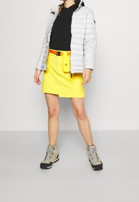 Giacca a vento imbottita grigio chiaro, felpa con cappuccio nera, gonna di denim gialla con tasca frontale e stivali da trekking beige con suole nere.