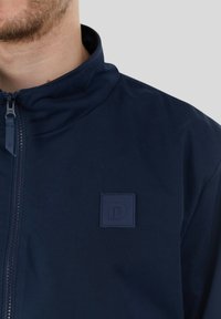 Marinblå zip-jacka med ståkrage, försedd med en texturerad logotypapplikation på bröstet, tillverkad av ett slätt, lätt tyg.