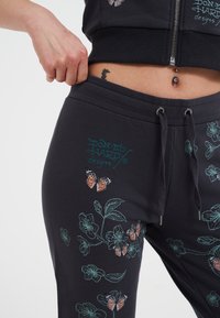 Mustat collegehousut, joissa on turkoosi ja oranssi kukka- ja perhoskirjonta, säädettävä nyöri ja pehmeä kuvio.