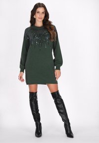 Grünes Strickkleid mit langen Ärmeln, das ein perlenbesetztes Design in vertikalen Linien am Ausschnitt aufweist. Kombiniert mit schwarzen Overknee-Stiefeln.