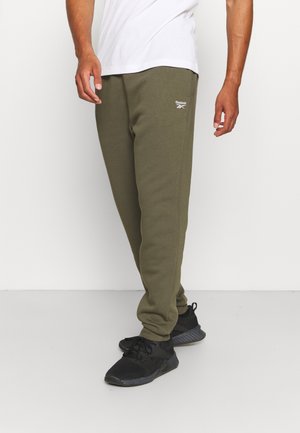 Mężczyzna w oliwkowozielonych spodniach dresowych Reebok, czarnych trampkach i białym t-shircie, stojący na tle jednolitego jasnego tła.