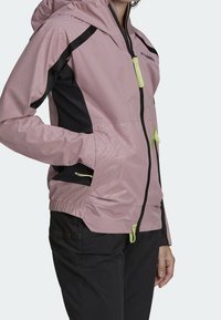 Ljusrosa vindjacka med svarta sidopaneler, dragkedja fram och gröna detaljer. Har en huva och elastiska ärmslut.