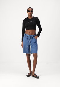 Čierny crop top s dlhým rukávom a logom, kombinovaný s modrými džínovými šortkami s elastickým pásom a rozstrapkaným okrajom. Čierne ploché topánky dotvárajú vzhľad.