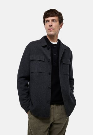 Veste en laine gris foncé avec deux poches poitrine, un col et un devant boutonné. Portée sur un pull en laine côtelé noir, associée à un pantalon en velours côtelé.