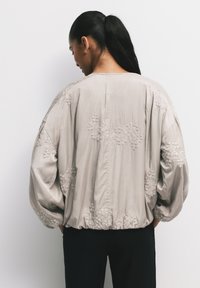 Blusa grigia chiara, dal taglio morbido con maniche voluminose e ricamo floreale. Tessuto testurizzato con orlo arricciato, caratterizzato da una lieve lucentezza.