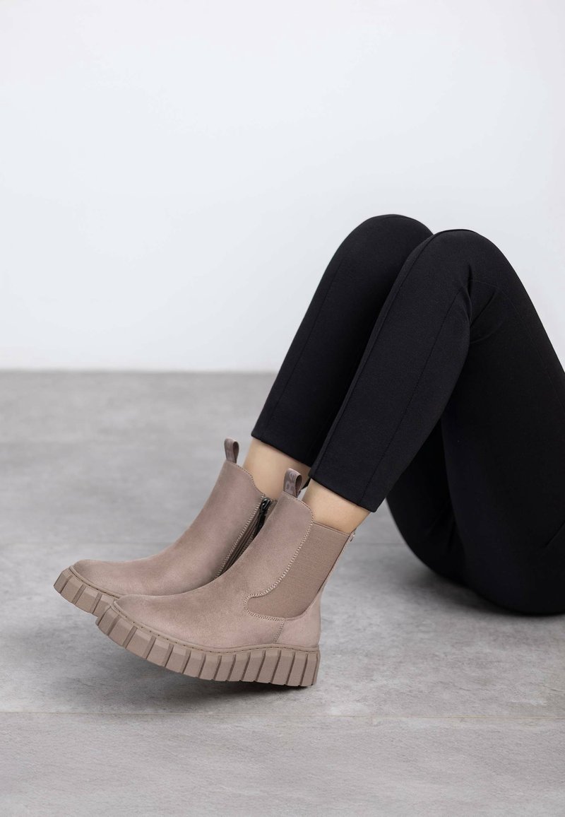 Personne portant des bottines en daim taupe avec des semelles épaisses et rainurées et un pantalon noir ajusté, assise jambes pliées sur un sol gris.