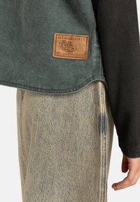 Groen T-shirt met korte mouwen en een bruine leren logopatch, gecombineerd met beige gescheurde spijkerbroek met zichtbare stiksels en textuur.