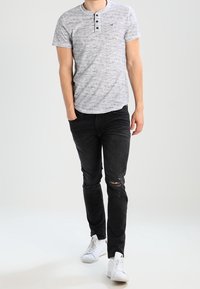 Camisa henley gris de manga corta con tres botones, jeans negros desgastados y zapatillas blancas. El conjunto presenta un diseño casual.