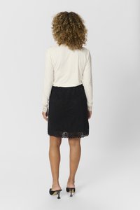 Fustă din dantelă neagră cu tiv ondulat, asociată cu un top crem cu mânecă lungă și ajustat. Ținuta include pantofi cu toc cu un design unic în formă de triunghi.