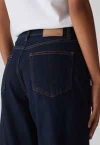 Personne portant un jean en denim bleu foncé avec un patch de marque en cuir tan et des coutures visibles, associé à un haut blanc.