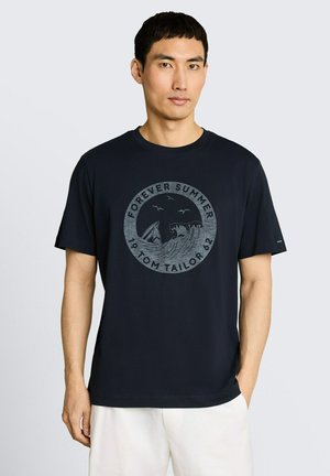 Mężczyzna w granatowej koszulce z okrągłym nadrukiem "Forever Summer" z falą i ptakiem oraz białych szortach, stojący na jednolitym tle.