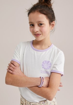 Jeune fille aux cheveux bruns attachés en chignon, portant un t-shirt blanc à bordure lavande avec le logo "Saffron Sunsets", les bras croisés et un léger sourire.