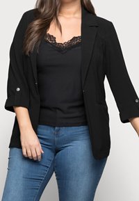 Zwarte blazer met opgerolde mouwen, sluiting met één knoop en zakken. Gemaakt over een zwarte top met kantkraag, gecombineerd met blauwe jeans.