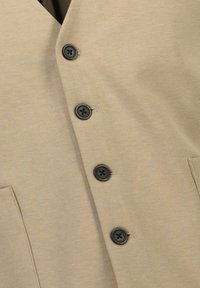 Beige skjorta med knappar som har fem svarta knappar, v-ringning och två framfickor. Texturen är mjuk med en subtil väv.